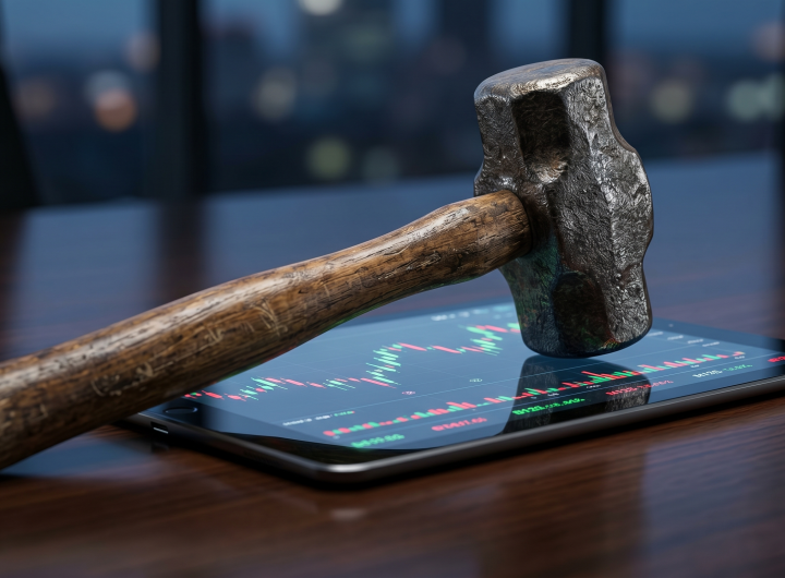 A heavy stone hammer resting on a tablet screen displaying a green and red stock market candlestick chart on a dark wooden desk.