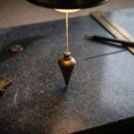 A brass plumb bob hanging over a granite drafting slab beside a brass square and a 24-inch gauge laid across a fresh pencil line.